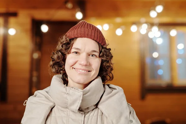 Happy young brunette woman with curly hair in warm winterwear looking at camera while standing against cottage decorated with lights