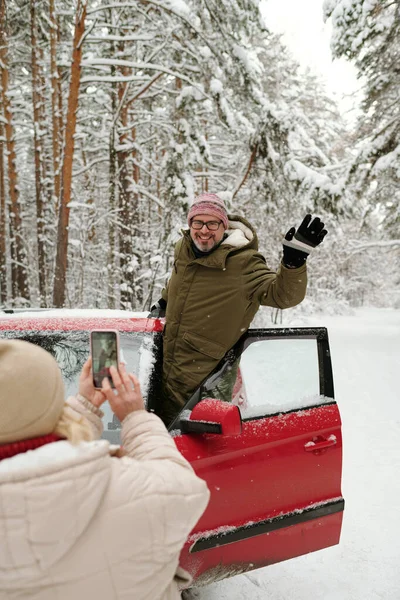 穿着冬衣的快乐而成熟的男性徒步旅行者一边站在红车门口一边挥手 一边用智能手机为妻子摆姿势 — 图库照片