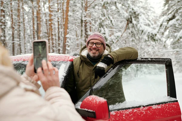 在森林里 戴着眼镜 穿着冬衣 快乐而成熟的男人站在敞开的车门边 为他的妻子摆姿势 拿着智能手机 — 图库照片