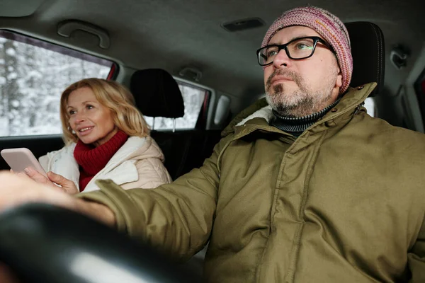 头戴眼镜 头戴便帽 头戴暖冬夹克的成熟男子 坐在妻子身边 开着汽车 手握方向盘 十分严肃 — 图库照片