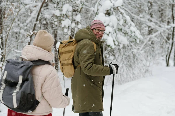 快乐而成熟的男人 背着背包和手杖 一边看着他的妻子 一边走在被积雪覆盖的林子里的路上 — 图库照片