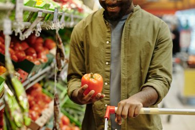 Süpermarkette sebzelerle sergilenirken elinde taze kırmızı biber tutan mutlu genç erkek tüketicinin yakın çekimi