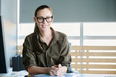 Formalite icabı yeni müşteriler beklerken masa başında oturmuş gülümseyen kameraya bakarken mutlu genç denetçi ya da İK yöneticisi.