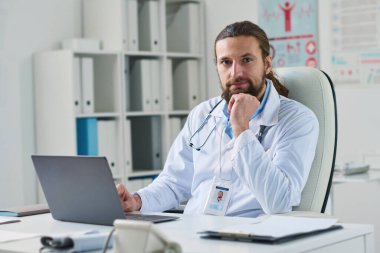 Laboratuvar önlüklü, genç ve başarılı bir klinisyen. İş yerinde bilgisayarının önünde oturuyor ve kameraya bakıyor.