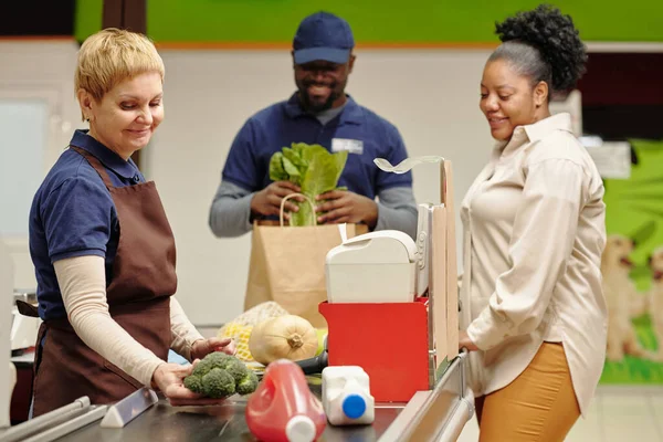 若いアフリカ系アメリカ人女性の消費者にサービスを提供しながら スーパーマーケットでチェックアウトカウンターに立って幸せな成熟したセールスマン — ストック写真