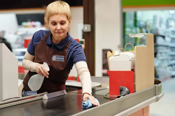 Mature female cashier in uniform spraying sanitizer on checkout line while bending over it and and wiping it with duster in supermarket