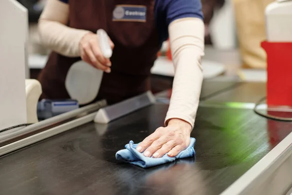 Close-up of cashier bending over checkout line in supermarket while spraying santizer on it and cleaning it before serving new customers