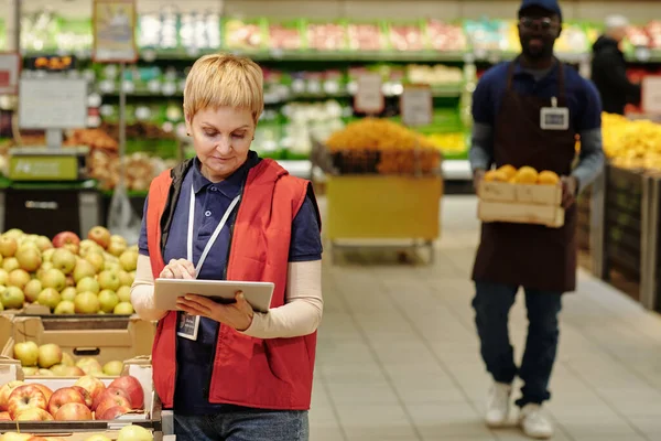 新鮮なリンゴと大きなディスプレイとリビジョン中にタブレットを使用して均一な立位のスーパーマーケットの熟女ブロンドの女性管理者 — ストック写真