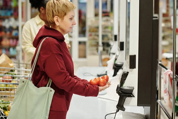 スーパーマーケットで製品の価格を示す画面の前に立って新鮮なトマトの山とブロンド成熟した女性の顧客の側面図 — ストック写真