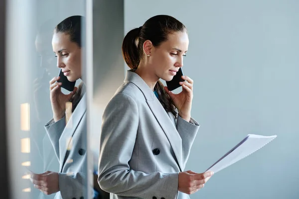 Young female financial director in grey suit looking through contract points and talking to business partner on mobile phone in office