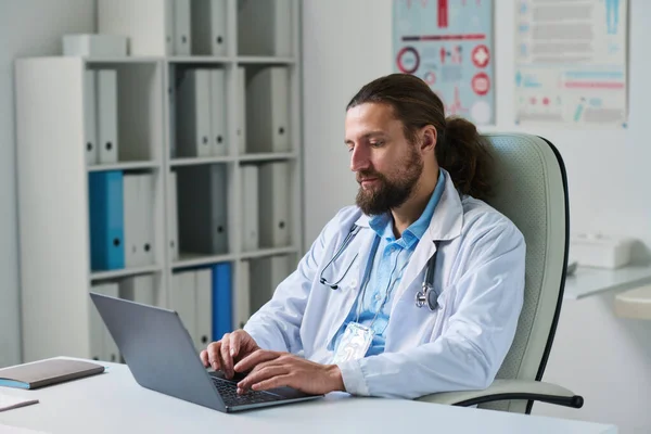 医療室の職場でオンライン患者に相談しながらノートパソコンのキーボードを入力する研究室のコートの若い深刻な医師 — ストック写真