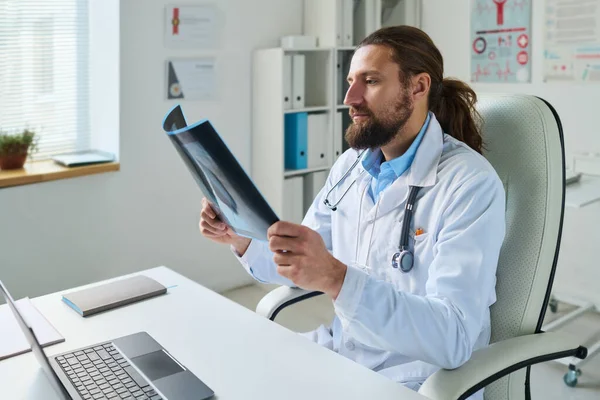 ラップトップの前の机でアームチェアに座ってオンライン患者を相談しながら X線結果を見て男性の放射線技師を自信を持って — ストック写真
