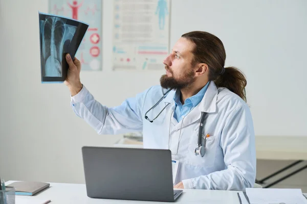 オンライン患者の肺のX線を注意深く見ている研究室のコートの男性臨床医は 相談中にノートパソコンの前で彼の職場に座っている間 — ストック写真