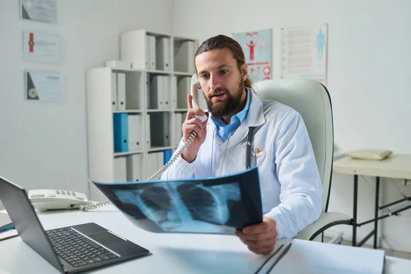 診療所の職場に座っている間に肺のX線を見て 電話受信機の患者と話すことを確信している放射線技師 — ストック写真