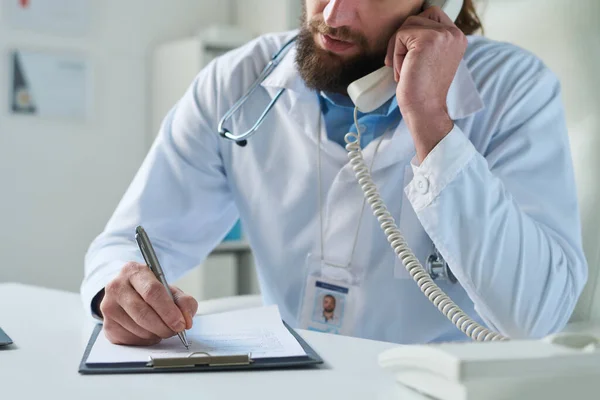 ノートを作成しながら クリップボードに医療文書の上にペンを保持し 電話で患者と話す研究室のコートの若い臨床医のクローズアップ — ストック写真