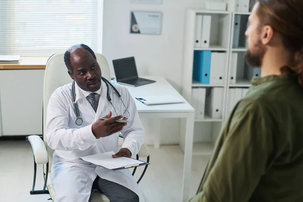 Pewny Siebie Dojrzały Lekarz Dokumentacją Medyczną Rozmawiający Młodym Pacjentem Siedzącym — Zdjęcie stockowe
