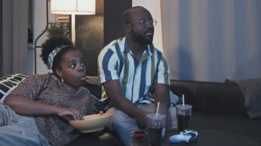 Orta uzunlukta bir Afro-Amerikan karı-koca kanepede dinleniyor, TV seyrediyor, abur cubur yiyor, içki içiyor, konuşuyor.