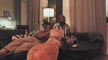 Afrika kökenli Amerikalı karı ve kocanın kanepede oturup televizyon izlemesi, abur cubur ve içki içmesi, konuşması, evcil köpekleri okşaması.