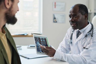 Genç hastaya tıbbi muayene sonuçlarını açıklayan kendinden emin bir radyolog tablet ekrandaki beyin taramasına işaret ediyor.
