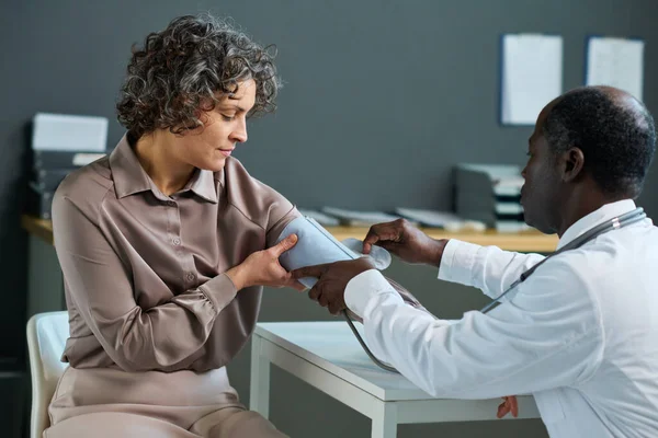 stock image African American male doctor using tonometer while sitting by workplace in hospital and measuring blood pressure of mature female patient