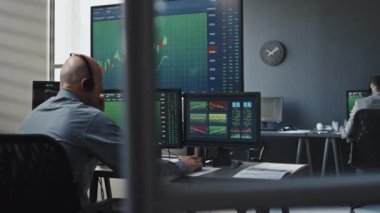Kulaklık takan profesyonel borsa simsarının bilgisayar monitörlerinde grafikler ve istatistiklerle çalışmasını ve müşteriyle bir şeyleri tartışmasını arka planda tut