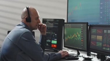 Kulaklık takan, mikrofonlu profesyonel borsa uzmanının bilgisayar monitörlerinin önünde müşteriyle stratejiyi tartışırken yan görüntüsü.