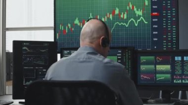 Kulaklık takan tanınmayan borsa simsarının bilgisayar monitörlerinin önünde veri ile çalışırken görüntüsü.