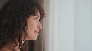 Pencere önünde dikilip perdelere dokunarak düğün merasimini düşünen, pullu bir elbise giyen modern beyaz gelinin yan görüntüsü.