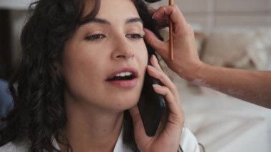 Uzun kıvırcık saçlı çekici gelin düğün hazırlıkları sırasında telefonda biriyle konuşuyor. Makyaj sanatçısı göz farı sürüyor.