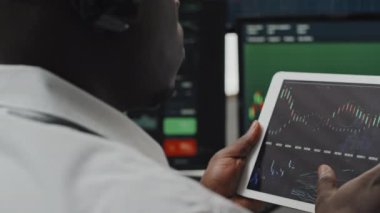 Kulaklık takan Afrikalı Amerikalı borsa simsarının dijital tablet ekrandaki döviz tablolarını izlediği ve müşteriyle dalgalanma sorunlarını tartıştığı yakın plan.