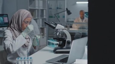 Koruyucu gözlüklü, eldivenli, test tüpündeki mavi sıvıya bakıp laboratuvar masasında oturan genç Müslüman kadın laboratuvar asistanı.
