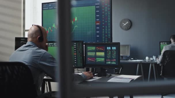 专业股票交易员头戴耳机在计算机显示器上与图表和数据一起工作并与客户讨论某事的后视镜 — 图库视频影像
