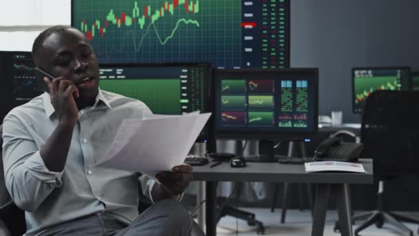 年轻的黑人股票交易者靠着桌子坐着 桌上拿着计算机显示器 屏幕上有货币统计数据 看文件中的数据 还在电话上交谈 — 图库视频影像