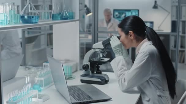 Mikroskop Kullanarak Modern Laboratuardaki Masada Oturan Siyah Saçlı Beyaz Elbiseli — Stok video