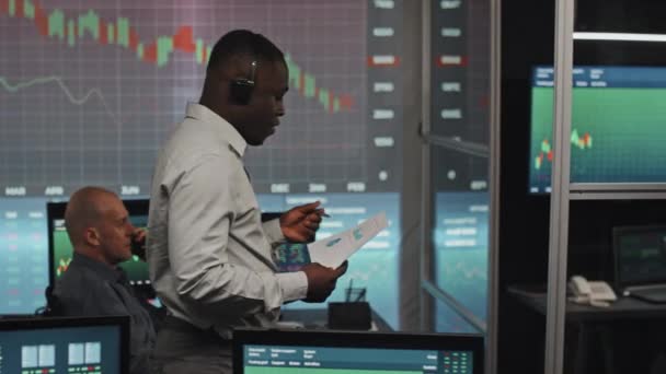 年轻的黑人股票交易顾问带着印有图表的耳机 深夜与客户通电话 他的高加索同事们正在做背景研究 — 图库视频影像