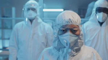 Laboratuvarda kameraya bakan bayan Asyalı doktor ve beyaz tehlikeli madde giysileri, solunum cihazları ve yüz kalkanlarının yakın plan portresi.