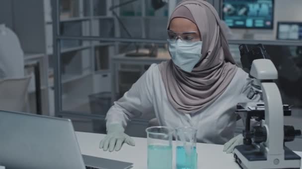 研究室の机に座っている白いガウン ゴーグルの若いイスラム教徒の女性研究室の技術者の胸 アップスローモーション 顕微鏡やポータブルコンピュータを使用して — ストック動画