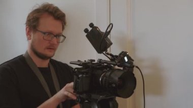 Gözlüklü genç adamın göğüs göğsü profesyonel video kamerayla çekim yapıyor ve içeride görünmez bir iş arkadaşıyla konuşuyor.