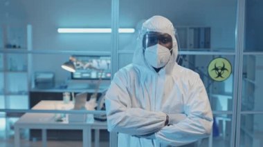 Beyaz tehlikeli madde giysisi, solunum cihazı ve yüz kalkanı giymiş, laboratuvarda kameraya bakan, kendine güvenen Afrikalı Amerikalı bir doktorun belden yukarı portresi.