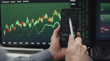 Tanımlanamayan olgun komisyoncu şirket uzmanı masasında bilgisayar monitörleriyle oturmuş akıllı telefon tutuyor ve veri tablolarıyla çalışıyor.