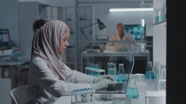 Laboratuvarda masada oturan, taşınabilir bilgisayarda yazan genç Müslüman kadının yan görüntüsü.