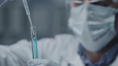 Bulanık eldivenli, gözlüklü ve yüz maskeli bilim adamının seçici odağı mavi sıvıyla test tüpüne bakıyor, ondan damlatıcıyla örnek alıyor.