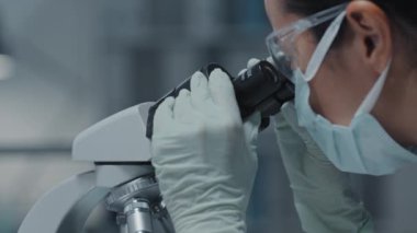 Mikroskop kullanarak koruyucu gözlük, eldiven ve maske takan genç bayan laboratuvar asistanının yan görüntüsü