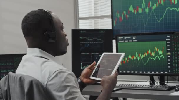 现代年轻的黑人股票交易专家坐在电脑屏幕前的办公桌前 手持数字平板电脑观看数据 并与客户通电话 — 图库视频影像