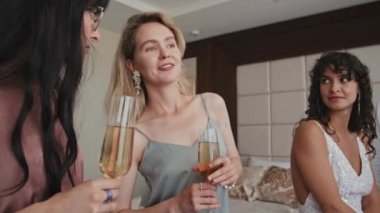 İki genç nedimenin elinde köpüklü şarap bardaklarıyla sohbet etmeleri, gelinin onlara bakması.