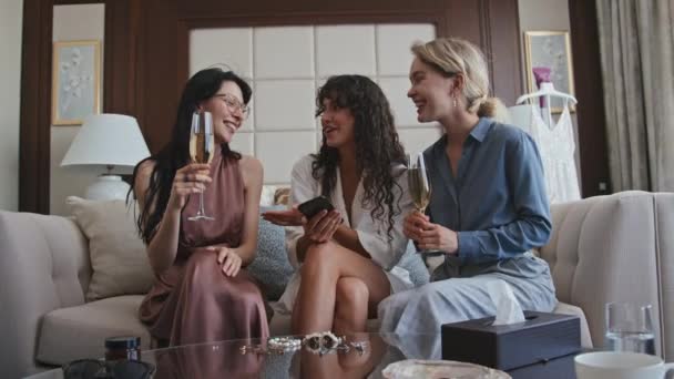 年轻的新娘和伴娘们坐在客厅的沙发上 手里拿着酒杯 手里拿着闪闪发光的葡萄酒 在智能手机上拍着自己的照片 — 图库视频影像