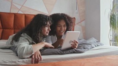 Genç Siyah ve Kafkasyalı kız arkadaşlar yatak odasında dinleniyor ve dijital tablette internette birlikte sörf yapıyorlar.