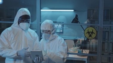 Koruyucu giysiler içinde laboratuvarda durup tablet bilgisayar kullanarak konuşan Asyalı ve Amerikalı kadın ve erkek doktorların orta solu.