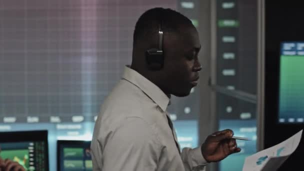 非洲裔美国经纪公司专家头戴耳机 手持文件在办公室走来走去 深夜通过电话讨论股票交易问题的画像 — 图库视频影像