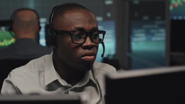 中程度のクローズアップOdアフリカ系アメリカ人男性を身に着けています眼鏡とヘッドフォンでマイク作業オフィスで夜遅くにコンピュータ上で — ストック動画